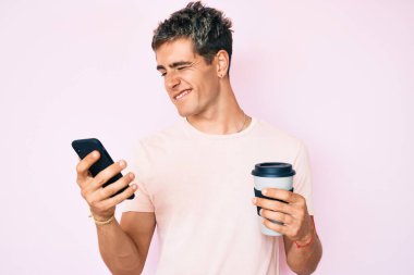 Akıllı telefon kullanan genç yakışıklı adam kahveyi alıp götürüyor kameraya seksi bir ifadeyle bakıyor, neşeli ve mutlu bir yüzle.. 
