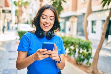 Genç İspanyol kadın şehirde akıllı telefon ve kulaklık kullanarak mutlu bir şekilde gülümsüyor..