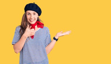 Fransız beresi takan ve çizgili tişört giyen genç ve güzel sarışın kadın avuç içini gösteriyor ve baş parmaklarıyla hareket ediyor, mutlu ve neşeli gülümsüyor. 