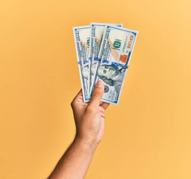 100 dolarlık banknotları izole edilmiş sarı arka planda tutan İspanyol adamın eli..
