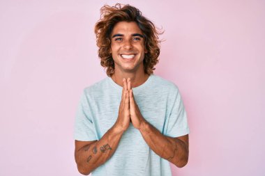 Gündelik kıyafetler giyen genç İspanyol adam elleriyle dua ediyor af diliyor kendinden emin bir şekilde gülümsüyor.. 