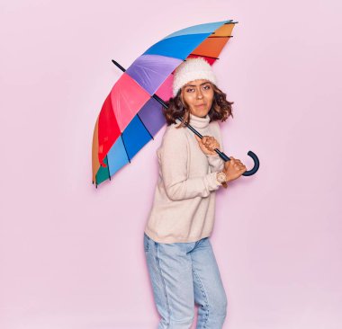 Şemsiye tutan genç, güzel Latin kadın mutlu gülümsüyor. İzole edilmiş pembe arka planda yüzünde gülümsemeyle zıplıyorsun.