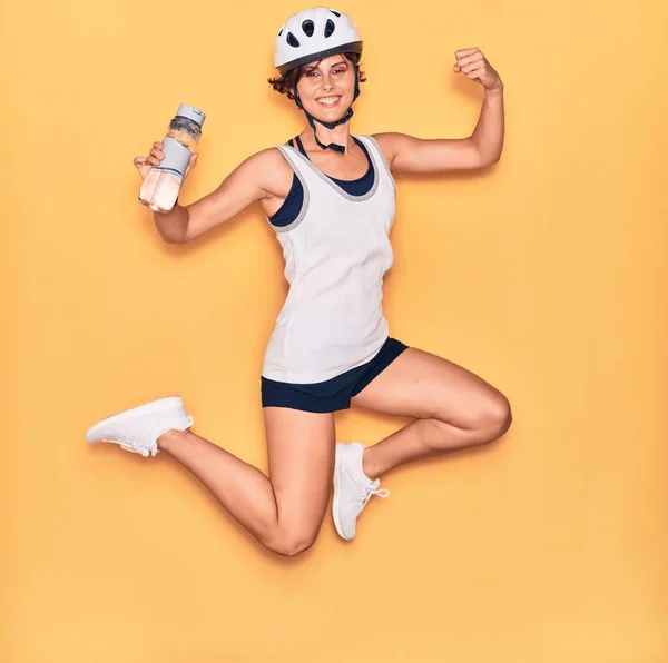 若い美しいバイカーの女性が幸せな笑顔自転車ヘルメットを着用 顔に笑顔でジャンプ隔離された黄色の背景に水のボトルを保持 — ストック写真