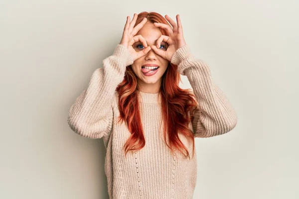 年轻美丽的红头发女人穿着宽松的冬季毛衣 像双筒望远镜一样做手势 伸出舌头 眼睛透过手指看过去 疯狂的表达 — 图库照片