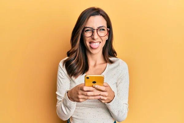 Młoda Brunetka Kobieta Pomocą Smartfona Żółtym Tle Wystający Język Szczęśliwy — Zdjęcie stockowe