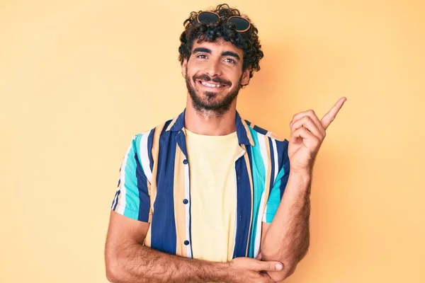 Handsome young man with curly hair and bear wearing summer shirt and sunglasses smiling happy pointing with hand and finger to the side