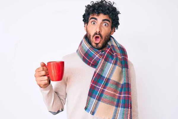 Handsome young man with curly hair and bear wearing winter scarf and drinking a cup of hot coffee scared and amazed with open mouth for surprise, disbelief face