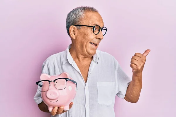 Przystojny Dojrzałe Senior Człowiek Gospodarstwa Świnka Bank Okulary Wskazując Kciuk — Zdjęcie stockowe