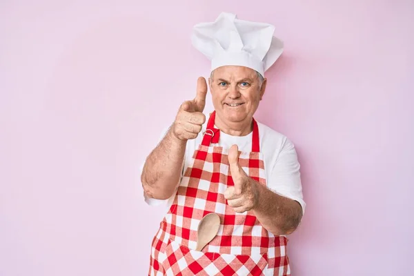 年长的白发男子穿着专业的面包师围裙 手指指向相机 脸上洋溢着快乐和滑稽的表情 充沛的精力和活力 — 图库照片
