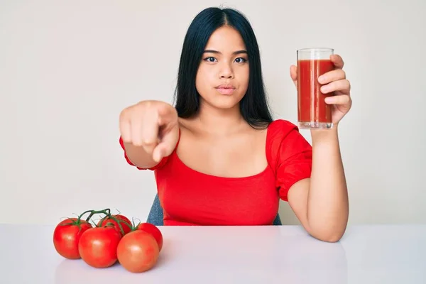 年轻美丽的亚洲女孩一边喝着一杯健康的番茄汁 一边用手指着相机和你 摆出一副严肃而自信的样子 — 图库照片