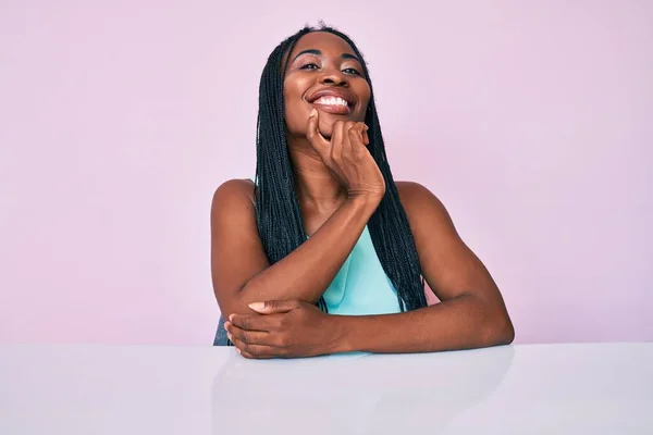 African american woman with braids wearing casual clothes sitting on the table looking confident at the camera with smile with crossed arms and hand raised on chin. thinking positive.