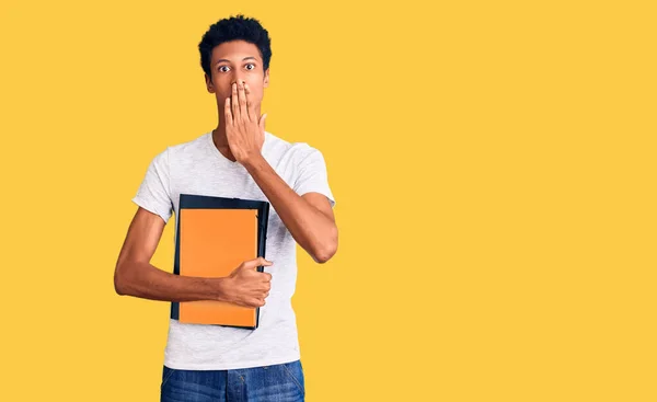 Joven Hombre Afroamericano Sosteniendo Libro Cubriendo Boca Con Mano Sorprendido —  Fotos de Stock