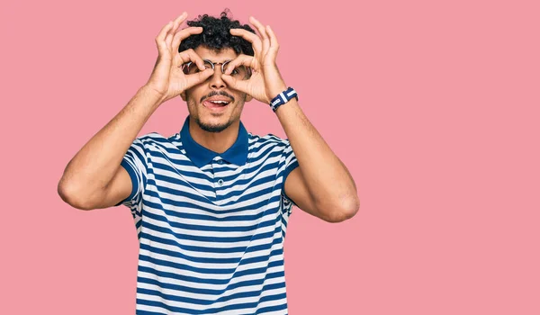 年轻的阿拉伯男子穿着休闲装 戴着眼镜 像双筒望远镜一样做手势 伸出舌头 眼睛透过手指看着 疯狂的表达 — 图库照片