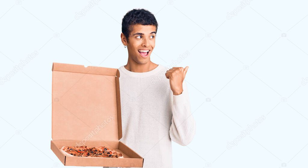 Young african amercian man holding delivery pizza box pointing thumb up to the side smiling happy with open mouth 