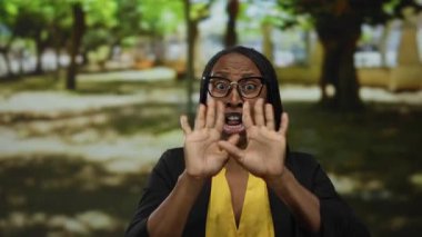Gözlüklü ve kıvırcık saçlı bir kadın, sarı gömlek ve siyah ceket giymiş, bulanık yeşil ağaçlar ve güneş ışığıyla çevrili park ortamında şaşırmış bir ifade sergiliyor..
