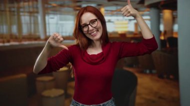 Kızıl saçlı ve gözlüklü bir kadın, kapalı bir restoran ortamında, hem onaylama hem de onaylamamazlık gösteren, gelişigüzel bir ortamda zıt duyguları vurgulayan baş parmak hareketleri yapar..