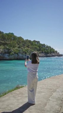 Santanyi 'deki güneşli plajda okyanus manzarasını yakalayan bir kadın, açık mavi gökyüzü altında canlı turkuaz su ve yemyeşil yemyeşil bir Mallorca..