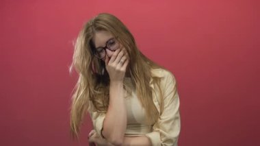Uzun saçlı ve gözlüklü genç sarışın kadın stüdyoda başını eğerken ağzını eliyle kapatıyor..