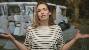 Ormanda ağaçların arasına park edilmiş hasarlı beyaz bir minibüsün önünde omuz silkerek el kaldıran bir kadın..