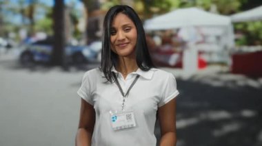 Dışarıda gülümseyen genç Latin bir kadın yumruğunu sallıyor ve arka planda bulanık bir şekilde güneşli bir sokakta kimlik kartıyla kamerayı işaret ediyor..