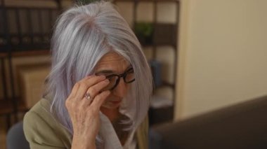 Ofisteki kıdemli kadın, gözlerini gözlüklerle ovalıyor, bir iş ortamında yorgunluk gösteriyor, raflarla çevrili ve modern kapalı bir ortamda bilgisayar kullanıyor..