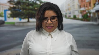 Arka planda ağaçlar ve binalar olan bir şehir caddesinde göz kırpan ve gülümseyen genç Latin bir kadın..