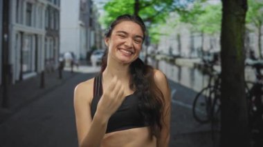 Latin kökenli genç bir kadın siyah spor sutyen giyip parmakla işaret parmağı kaldırarak kentteki kaldırım taşı sokağına bakıyor..