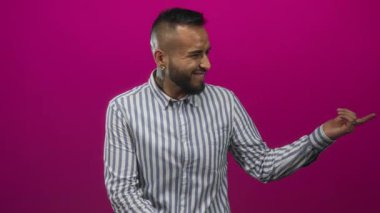 Çizgili tişörtlü, sakallı ve kulağında piercingler olan bir adam işaret eden elle gülümsüyor ve eflatun bir stüdyoda gözleri kapalı; neşe.