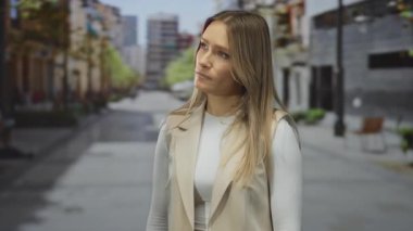 Şehir caddesinde dikilen sarışın bir kadın, beyaz bir üst ve bej bir yelek giyiyor, şehir içinde düşünceli yüz ifadesini vurguluyor..