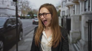 Genç kadın, şehir caddesinde park halindeki arabaların yanında ağzı açık gülümsüyor; keyif iyimserliği.