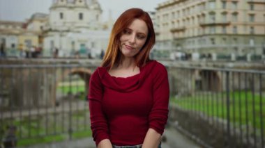 Şehir caddesinde kızıl saçlı bir kadın kırmızı bir kazak giyiyor. Gündüz vakti bulanık şehir mimarisine karşı düşünceli görünüyor.