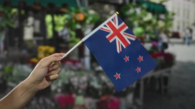Yeni Zelanda bayrağını şehir caddesindeki çiçekçinin yakınında tutarak canlı şehir atmosferini ve ulusal gururu yakalıyor..