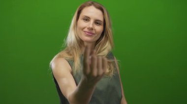 Genç sarışın kadın, parmaklarıyla canlı yeşil izole bir arka plana doğru geliyor. Sıcak bir gülümsemeyle dostça bir davet sunuyor..