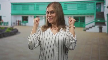 Sarışın kadın yumruklarını sıkıyor ve güneş ışığı altında yeşil bir binanın önünde gülümsüyor..