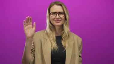 Gülümseyen ve parlak pembe arka plana el sallayan gözlüklü ve spor ceketli bir kadın stüdyo ortamında dostça bir jest yapıyor..