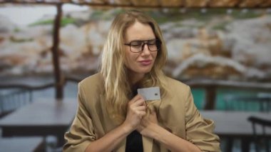 Şehir terasında elinde kredi kartıyla kaygı gösteren genç sarışın bir kadın açık hava kentsel restoranındaki finansal stresi yansıtıyor..
