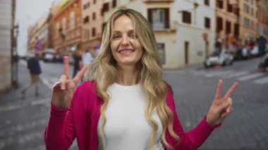 Sarı saçlı, pembe kazaklı, hareketli şehir caddesinde gülümseyen kadın gündüz vakti açık havada barış işareti yapıyor ve şehir yaşamını ve mutluluğunu gözler önüne seriyor..