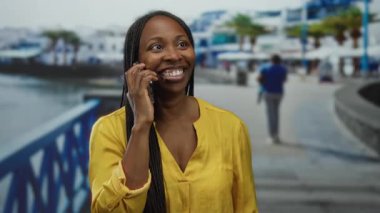 Açık hava güneşli deniz kenarında akıllı telefondan konuşan bir kadın. Arka planı bulanık sarı gömlekli, plajı ve palmiye ağaçlarını gösteren rahat bir ortam yakalayan bir kadın..