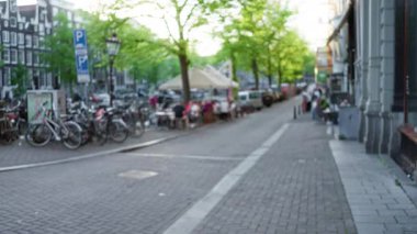 Amsterdam 'da bisikletli bulanık sokak manzarası, dışarıda yemek yiyen insanlar, ve temiz bir gökyüzünün altında kaldırımı kaplayan canlı yeşil ağaçlar..