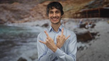 Elleri çapraz duran gülümseyen adam okyanus kıyısında altın güneş ışığı altında kaya boynuzları yapıyor; rock metali eğlenceli.