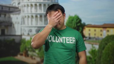 Asyalı, yeşil gönüllü gömlekli, İtalya 'da sidik kulesinin önünde duran açık hava ortamında düşünceli bir duyguyu ifade eden, arka planda ikonik bir simge olan.