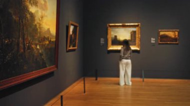 Müzede bir kadın, duvarlarda çerçevelenmiş resimlere bakıyor. Kapalı alanda ahşap zemin ve bulanık odaklı sanat galerisi..