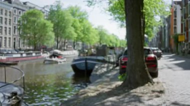 Amsterdam kanalı manzarası, bulanık ağaçlarla kaplı cadde, suda kayıklar, ve güneş ışığı çözülmüş bokeh etkisi yaratıyor, Hollanda 'nın sakin kentsel manzarasını yakalıyor..