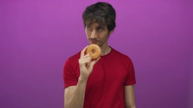 Kırmızı tişörtlü bir adam bir eliyle jöleli donut tutuyor ve diğer eliyle mor stüdyoda tamam işareti yapıyor..