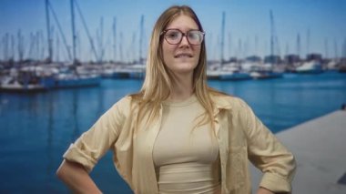 Gözlüklü sarışın kadın mavi suyun üzerinde güneşli bir iskelede teknelerin yanında burnunu çimdikliyor..