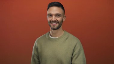 Süveter giyen genç İspanyol bir adam canlı turuncu bir arka plana karşı dostça bir gülümsemeyle beni arar, pozitif iletişim ve modern tarz sergiler..