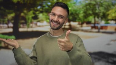 Güneşli bir parkta gülümseyen İspanyol bir adam. Arka planda ağaçlar ve yeşillik olan başparmaklarını gösteriyor. Sıradan giyinmiş, pozitif enerji yayıyor.