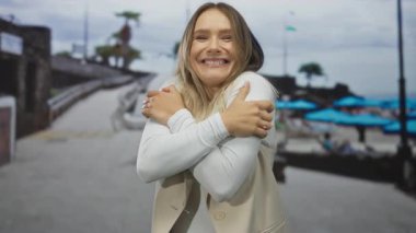 Deniz kenarındaki bir yerde gülümseyen ve kendini kucaklayan bir kadın, bulanık arka planda mavi şemsiyeler ve plaj ortamı rahatlamayı öneriyor..