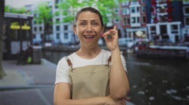 Bej elbiseli kadın geniş bir şekilde gülümsüyor ve binaların oradaki sokak kanalında elleriyle parmaklarını çimdikliyor;.