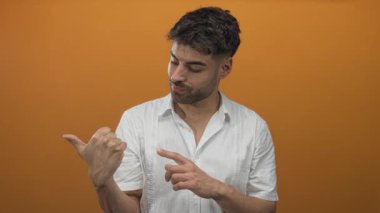 Adam, turuncu stüdyo arka planda beyaz gömlek giyerek, el ele bakarak çağrı hareketi yapıyor; davet.